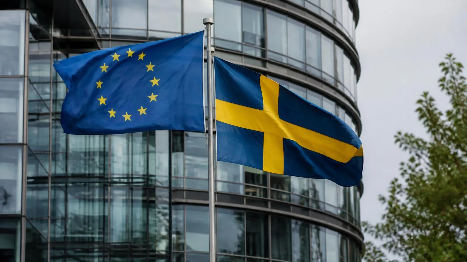 EU-flaggan och den svenska flaggan vajar framför en modern glasbyggnad i dagsljus